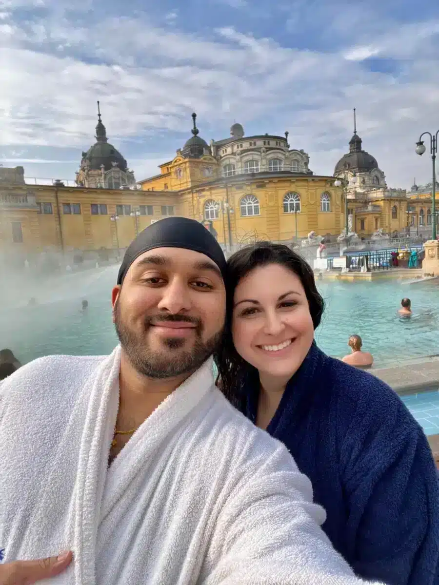 Paar-Selfie in Bademänteln vor dem dampfenden Außenbecken des Széchenyi Thermalbads Budapest im Winter