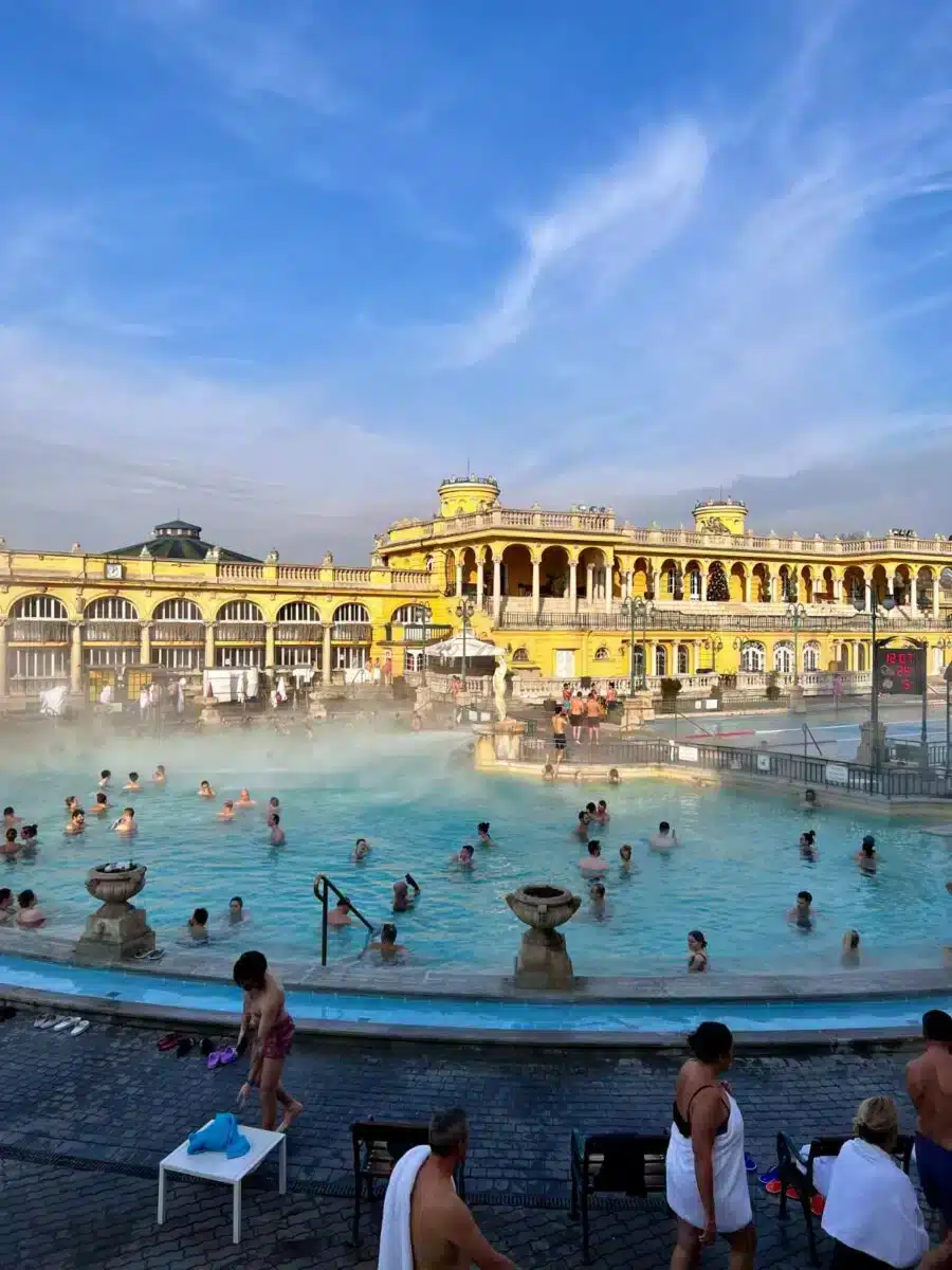 Belebtes Außenbecken des Széchenyi Thermalbads Budapest mit Badegästen und aufsteigendem Dampf vor der gelben Fassade