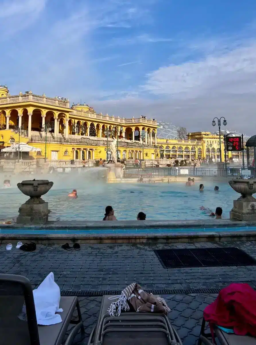 Blick von der Liegewiese auf das dampfende Thermalbecken und die gelbe Fassade des Széchenyi Bads Budapest