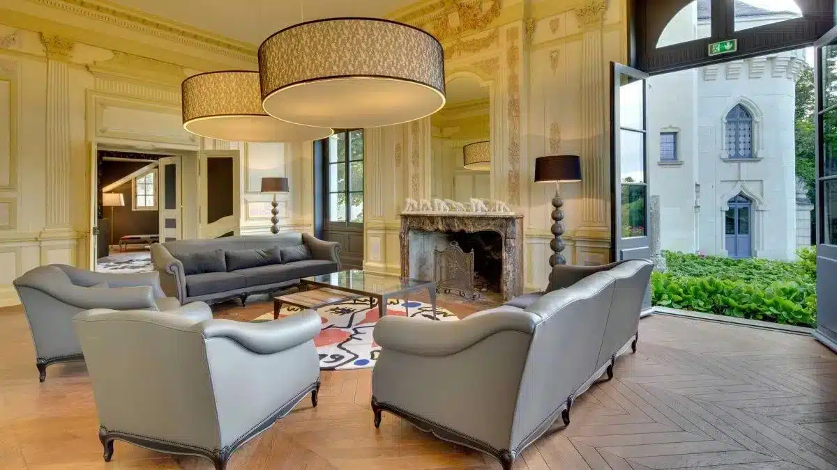 Großer Salon im Château de Candes im Loire-Tal mit prachtvollem Steinkamin, grauen Polstersesseln, Fischgrätparkett und großer Pendelleuchte