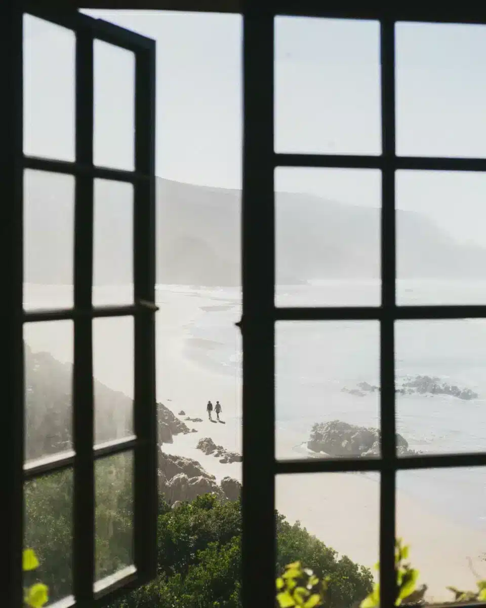 Blick durch dunkle Fensterscheiben auf eine neblige Küstenlandschaft mit zwei Silhouetten, die an einem abgelegenen Strand entlanggehen, fotografiert in einer Insider Villas Unterkunft