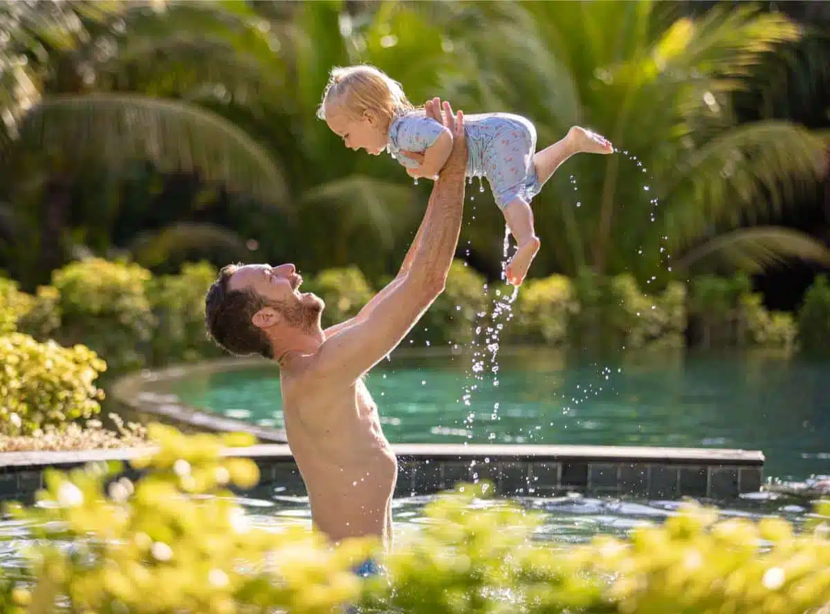 Vater hebt ein lachendes Kleinkind freudig über das Wasser in einem tropischen Villenpool, umgeben von üppigen Palmen, aufgenommen in einer Insider Villas Unterkunft