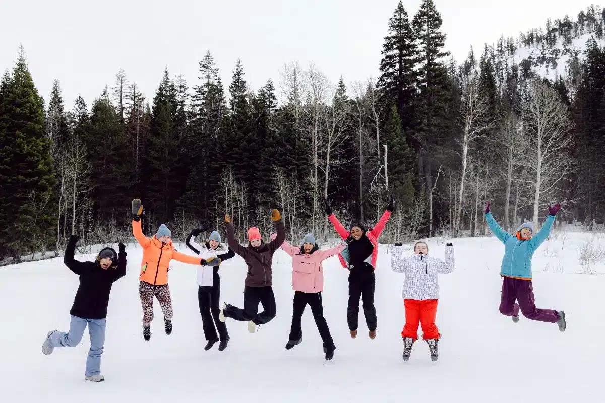Group of women jumping in the snow wearing colorful ski gear with pine trees in the background at a Retreat Company ski retreat