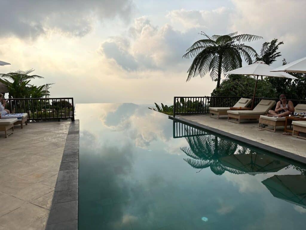 Infinity-Pool der Munduk Moding Plantation Bali in der Abenddämmerung mit dramatischem Himmel, der sich im stillen Wasser spiegelt, und Liegen am Rand