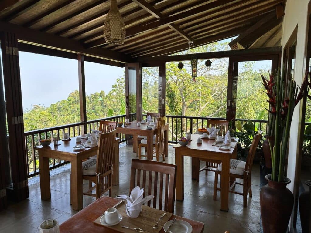 Offene Speiselounge der Munduk Moding Plantation Bali mit Teakholztischen und -stühlen mit Blick über das tropische Hochland-Blätterdach