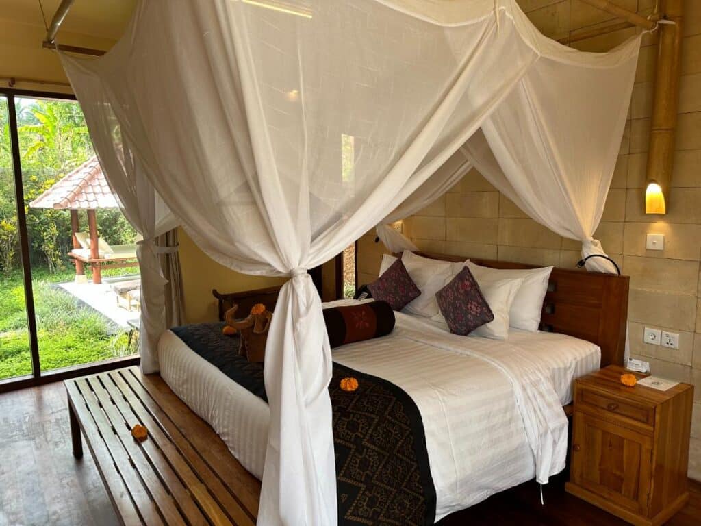 Weißes Himmelbett mit transparenten Vorhängen in der Munduk Moding Plantation Villa mit raumhohen Fenstern und Blick auf den tropischen Garten