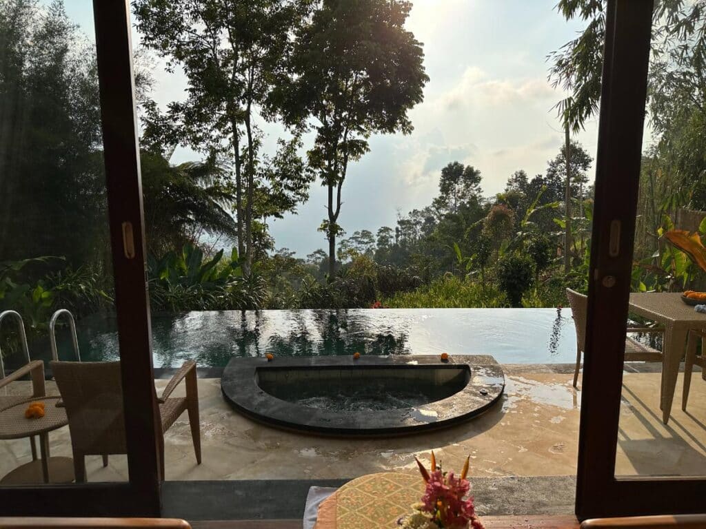 Blick auf den privaten Villenpool und Whirlpool der Munduk Moding Plantation Bali durch offene Holztüren, eingerahmt von hohen Bäumen
