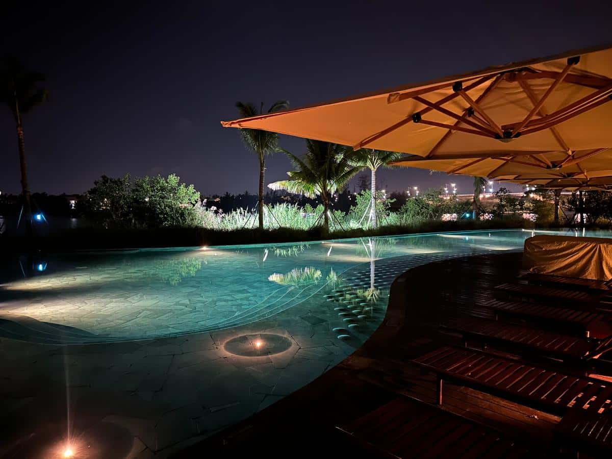 Main pool lit at night with palm reflections and timber loungers, Namia River Retreat, Hoi An, Vietnam